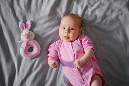 Smiling newborn girl in a pink onesie sticking out her tongue, lying next to a plush bunny rattle,...