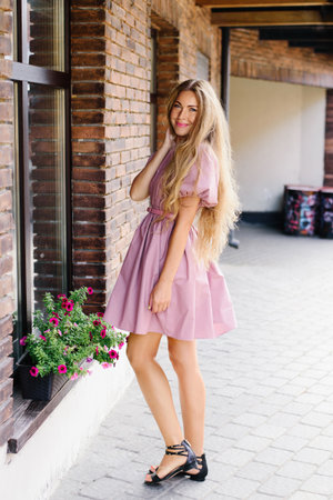 Young woman in a pink summer dress standing by a brick wall with flowers, ideal for lifestyle, fashion, and seasonal photography.の写真素材