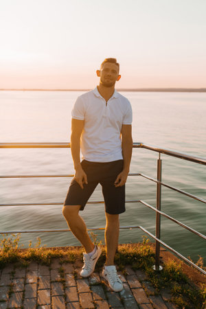 Stylish young man in a white polo shirt and dark shorts standing by the waterfront at sunset, posing casually against a metal railing. Perfect for lifestyle, fashion, travel, and summer leisure concepts for advertising and editorial use.の写真素材