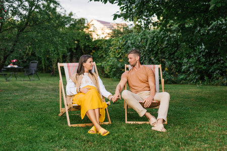 Smiling pregnant woman and her husband relaxing outdoors in chairs, holding hands and enjoying peaceful family time together.の写真素材