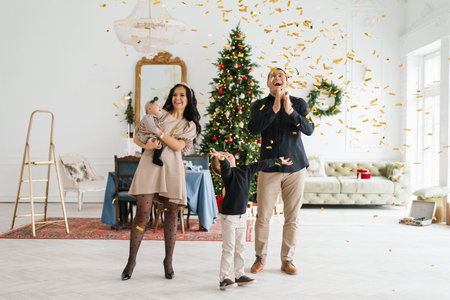 Cheerful family with two children celebrating Christmas at home, throwing golden confetti near a...