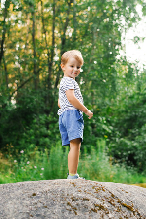 A smiling boy standing confidently on a large rock in a summer forest, surrounded by greenery and sunlight.の写真素材
