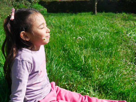 Relaxed little girl sitting in the grass in the sunshine, holding a flower in his mouthの写真素材