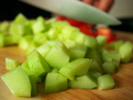 Close-up of a chopped cucumber with a man slicing a tomato in the background. A man is preparing a recipe with fresh summer vegetablesの写真素材