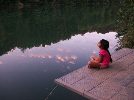 Little girl sitting on a pontoon by the river and contemplating the sky of a sunsetの写真素材