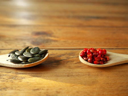 Two wooden spoons filled with pink peppercorn and pumpkin seeds on wooden backgroundの写真素材