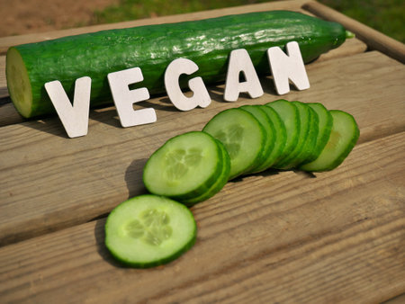 A closeup shot of ripe fresh sliced cucumber and Vegan word from wooden letters on a cutting boardの写真素材