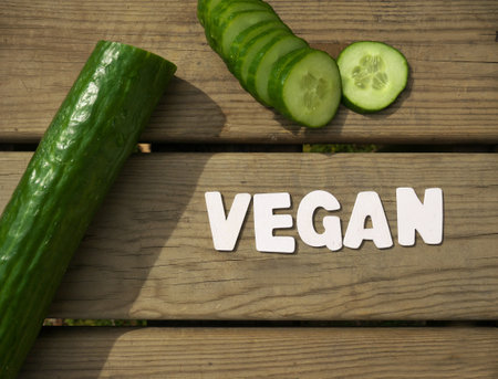 A top view of ripe fresh sliced cucumber and Vegan word from wooden letters on a cutting boardの写真素材