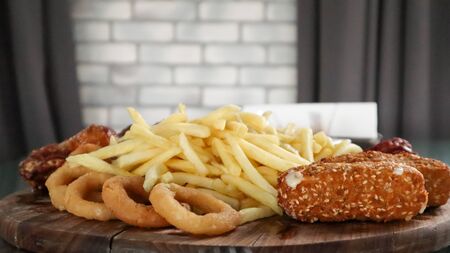 Wooden board with french fries, cheese sticks, onion rings, fried chicken wings, smoked sausages with sauce. Pub snacks menu. A high-calorie snack for group friends. Tasty snacks. Beer snackの写真素材