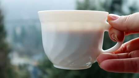 Girl hand holds a cup of white coffee and has a blurred green nature background. Mug in a female...