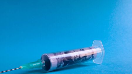 A roll of 100 dollars money inserted into a white medical hypodermic needle on a blue background. The concept of health care costs, credit, investment. Copy space for your text or logoの写真素材