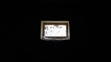 a small rectangular dirty and dusty web window in a large basement. White light penetrates the dark basementの写真素材