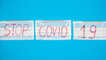 Typical 3-ply white surgical mask with rubber earhooks to cover mouth and nose with English block letters Stop Covid-19 on a blue background. Dangerous virus and quarantine concept. Copy space.の写真素材
