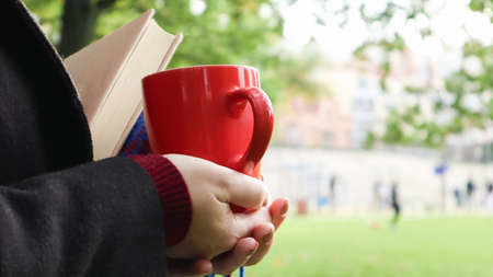 A red cup of coffee and a book with a blue checkered woolen blanket or plaid in the hands of a woman wearing a sweater and a black coat in the park. Warm and sunny weather. Soft cozy photography.の写真素材