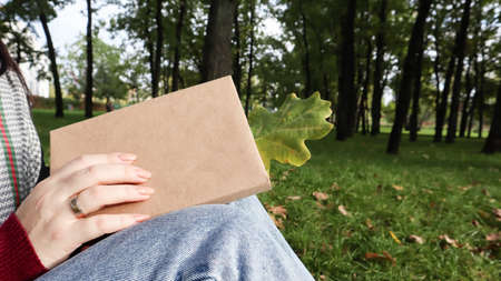 A woman holds a closed book lying in her lap with a fallen oak leaf close-up in a park on a sunny...