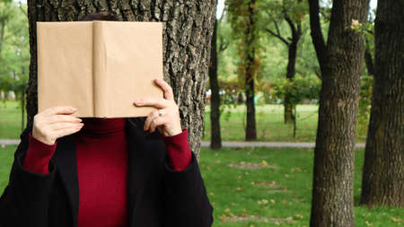 Portrait of a young woman with an open untitled book held close to her face. Brunette covering her...