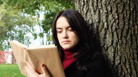 Portrait of a beautiful young woman who is sitting under a tree and reading her favorite book in a...