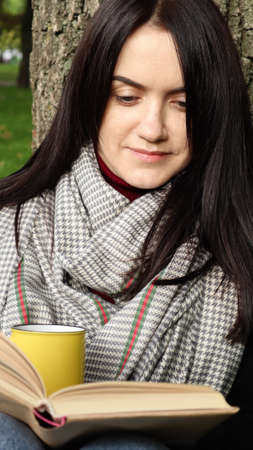 A woman sits under a tree and reads her favorite book while drinking coffee or tea from a yellow cup...
