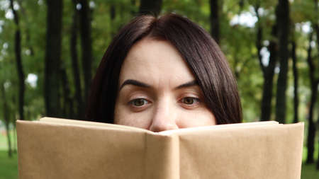 Portrait of a young woman with an open untitled book held close to her face. Brunette covering half...
