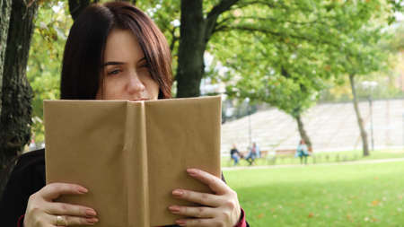 Portrait of a young woman with an open untitled book held close to her face. Brunette covering half...