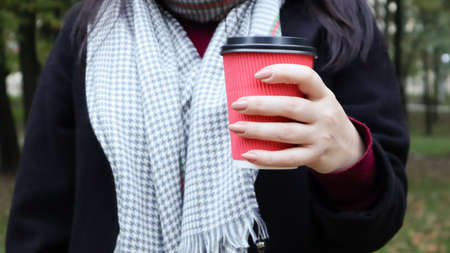 A young girl in a coat holds morning coffee with her while walking in the park. Hand holding paper cup of coffee in green park. Takeaway cappuccino. Close-up.の写真素材