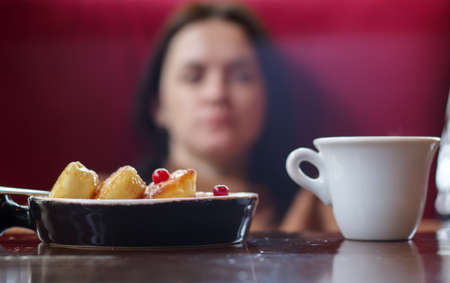 Portion of round paneer cheesecakes on a plate on a table with a white cup of coffee on a blurred background of an unrecognizable girl and restaurant interior. Healthy morning breakfastの写真素材
