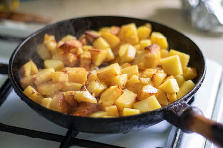 Roasting fresh potatoes in a cast iron skillet with sunflower oil. A view of a stovetop with a frying pan filled with golden fried potatoes in a real kitchen. food cookedの写真素材