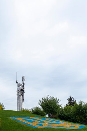 Statue of the Motherland against the blue sky. Coat of arms of Ukraine on the lawn, lined with blue and yellow stones. The trident, the official symbol of the state. Ukraine, Kyiv - October 08, 2022のeditorial素材
