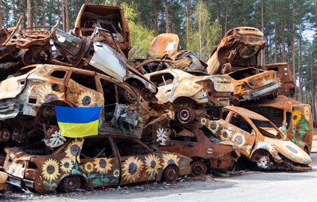 The national flag of Ukraine against the background of ruins and rusty metal of destroyed cars. A blue-yellow flag waving in the wind under the sun in a ruined city. Ukraine, Kyiv - April 29, 2023.のeditorial素材