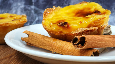 One Pastel de nata or Portuguese egg tart and cinnamon sticks on a white plate. Pastel de Belm is a...