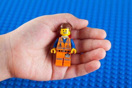 Tambov, Russian Federation - July 26, 2014 LEGO Hard Hat Emmet minifigure in child hand against Lego blue baseplate background. Studio shot.のeditorial素材