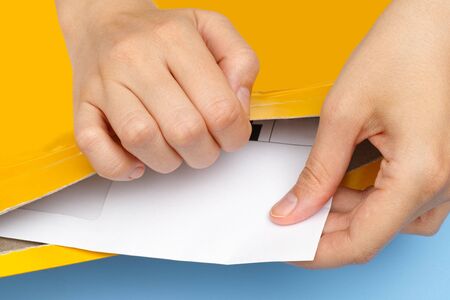 Woman hands getting out white envelope from yellow package. Blue background.の写真素材