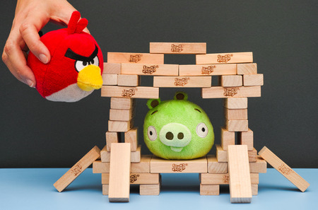 Tambov, Russian Federation - March 03, 2016 Bad Piggy sitting inside of house, made from Jenga bricks. Red Angry Bird in woman hand flying to broke Jenga house. Studio shot.のeditorial素材