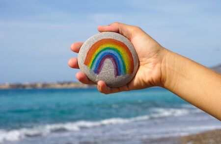 Woman hand hold pebble with painted rainbow against blue sky and sea.の写真素材