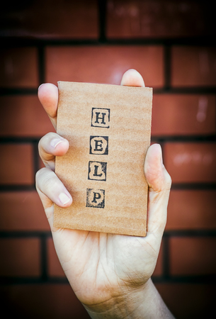 Woman hand holding cardboard card with word HELP made by black alphabet stamps against brick wall background.の写真素材