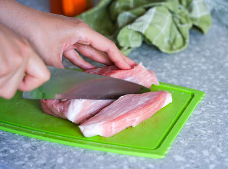 A woman with a knife cutting a raw pork on a green cutting board on a kitchen table. Some pepper and salt are scattered aroundの写真素材