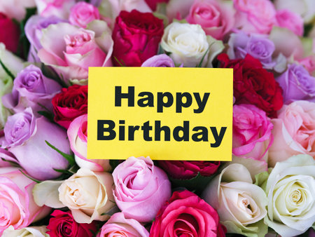 A yellow greeting card with words Happy Birthday laying on a big bouquet of colorful rosesの写真素材