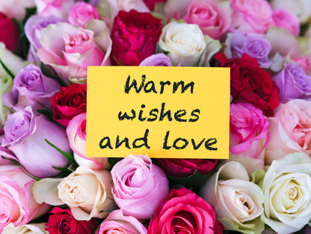 A yellow greeting card with words Warm Wishes and Love laying on a big bouquet of colorful rosesの写真素材