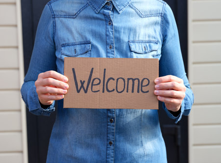 Woman holding a piece of cardboard with word Welcome and standing against the door of a house.の写真素材