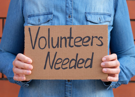 Woman holding piece of cardboard with words Volunteers Needed against brick wall background.の写真素材