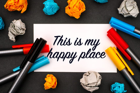 White paper blank with words This is My Happy Place, colour pens and crumpled paper balls on a black...