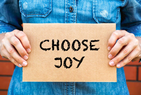 Woman holding a piece of cardboard with words Choose Joy against brick wall background.