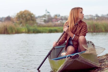 Young woman sitting in boat on autumn day against dull sky and town view landscapeの写真素材