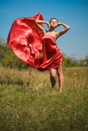 Young beautiful woman in red dress flying in wind against blue sky backgroundの写真素材