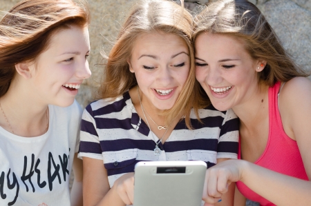 Three happy teen girl friends looking on tablet pc while sitting on green lawn on summer dayの写真素材