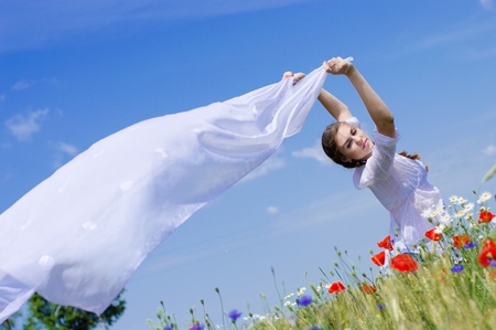 Young smiling woman standing in yellow wheat and poppy field holding a white long piece of cloth in the wind.の写真素材