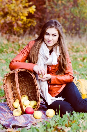Happy young woman with basket of fresh apples sitting on meadow on bright autumn dayの写真素材