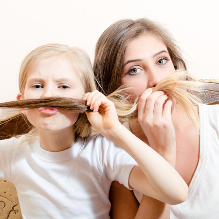 having fun making mustache beautiful young woman and little blond girl dressed in white shirts sitting on sofa & looking at camera close up portrait image on white copy space background above themの写真素材