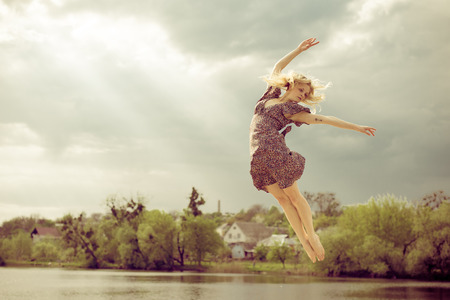 joyful freedom: portrait of beautiful blond dancing young woman with falling sun light rays from blue sky at river or water lake on spring or summer nature green outdoors background copy space pictureの写真素材