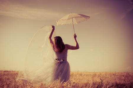 alone in the field: elegant romantic woman in long white dress having fun holding up parasol standing back to camera on blue sky copy space backgroundの写真素材